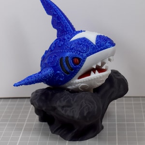 May include: A 3D-printed shark figurine with a blue body, white accents, and red eyes. The shark has intricate floral patterns and is mounted on a dark gray, rock-like base. The shark's mouth is open, revealing white teeth and a red interior.