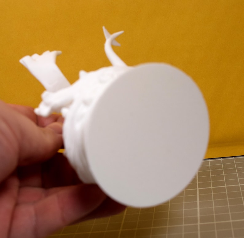 May include: A white, unpainted 3D printed figurine featuring a circular base and a sculpted creature with a pointed tail and textured details. The object is shown from an underside angle, highlighting the smooth, flat, circular base surface.