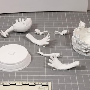 May include: A collection of white 3D printed model parts including a circular base, a torso, two hands, two small decorative pieces, and a textured wave-like structure. A ruler at the bottom shows scale, and a stack of colorful plastic bricks is visible.