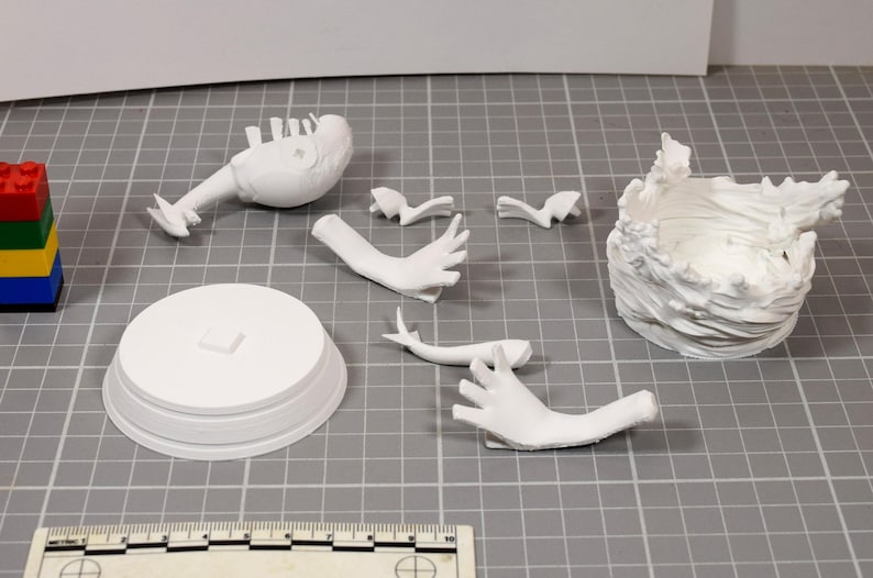 May include: A collection of white 3D printed model parts including a circular base, a torso, two hands, two small decorative pieces, and a textured wave-like structure. A ruler at the bottom shows scale, and a stack of colorful plastic bricks is visible.