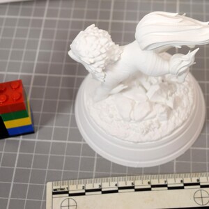 May include: A white unpainted figurine of a winged creature with a textured mane and tail, mounted on a circular base with rocky detailing. A ruler showing metric measurements is positioned at the bottom, and a stack of colorful plastic blocks is nearby.