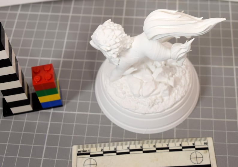 May include: A white unpainted figurine of a winged creature with a textured mane and tail, mounted on a circular base with rocky detailing. A ruler showing metric measurements is positioned at the bottom, and a stack of colorful plastic blocks is nearby.