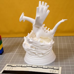 May include: A white 3D printed figurine of a dragon-like creature with large wings and a long tail, standing on a base sculpted to resemble swirling water. A ruler is visible at the bottom, showing a scale of 10 centimeters for size reference.