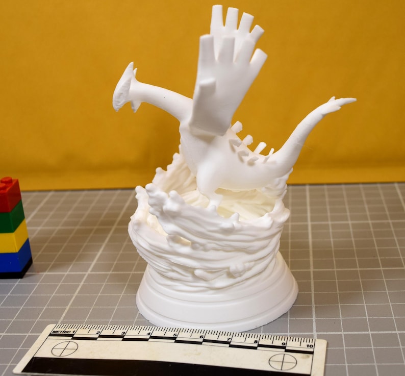 May include: A white 3D printed figurine of a dragon-like creature with large wings and a long tail, standing on a base sculpted to resemble swirling water. A ruler is visible at the bottom, showing a scale of 10 centimeters for size reference.