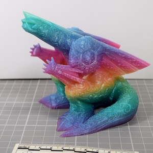 May include: A colorful, rainbow-hued fantasy creature figurine. The figurine has a detailed, textured surface with intricate patterns. The creature has wings, clawed feet, and a long tail. The color transitions from blue to pink to yellow and green.