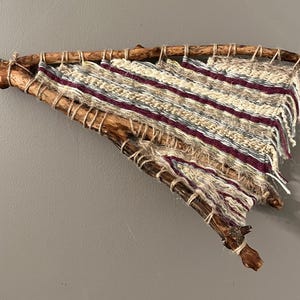 May include: A triangular wall hanging featuring a woven textile stretched across two intersecting natural wooden branches. The fabric displays horizontal stripes in cream, deep burgundy, and grey, with frayed edges and twine lashing.