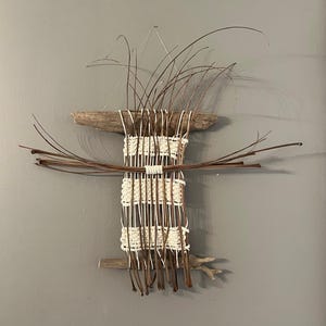May include: A handcrafted wall hanging featuring a woven design with natural elements. The artwork incorporates driftwood, cream-colored woven sections, and dried brown grasses, creating a rustic and organic aesthetic. The piece is mounted on a gray wall.