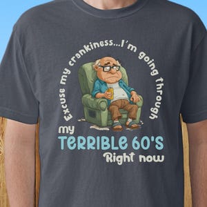 May include: Dark grey t-shirt with the text "Excuse my crankiness...I'm going through my TERRIBLE 60'S Right now." The graphic shows an elderly person in a chair. The word "pepper" is in the bottom left corner.