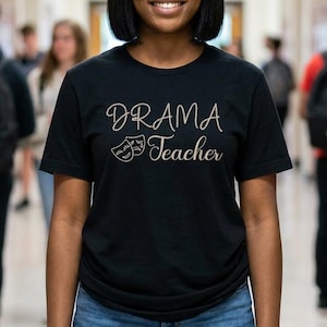 May include: A black t-shirt with the words "DRAMA Teacher" in a cursive font, along with two theater masks. The person is wearing blue jeans and is standing in a school hallway. Other students are in the background.