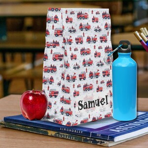 May include: A white lunch bag with a pattern of red fire trucks and firefighters, personalized with the name "Samuel". A bright red apple and a blue water bottle sit next to the bag on top of a stack of books.