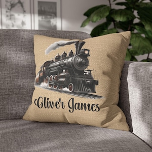 May include: Tan square throw pillow featuring a detailed illustration of a black steam train with white smoke. The text "Oliver James" is printed in a script font below the train. The pillow is on a gray sofa.
