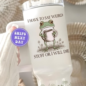 May include: White tumbler with a clear lid featuring a cartoon frog holding a coffee mug. The text reads "I HAVE TO SAY WEIRD STUFF OR I WILL DIE". The frog is green with brown eyes, and the mug is light pink.