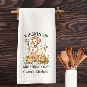May include: White kitchen towel with a cartoon illustration of a duck wearing a pink apron and holding a whisk. The duck is standing next to a bowl of batter and a cat sitting on a stool. The text "Whisk'n Up Something Cozy Gwen's Kitchen" is printed on the towel.