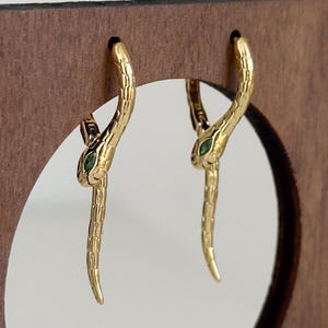 May include: A pair of gold-coloured snake earrings with green gemstone eyes. The earrings are displayed on a wooden stand with a circular cutout. The snake earrings have a textured design and are hanging from the stand.