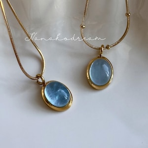 Peut inclure: Deux colliers dor&eacute;s avec des pendentifs ovales bleu clair. Les pendentifs sont sertis dans des montures dor&eacute;es. Les colliers sont pr&eacute;sent&eacute;s sur une surface blanche textur&eacute;e. Le texte "Nanahodream" est &eacute;crit en cursive.