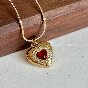 May include: Gold-toned necklace featuring a heart-shaped pendant. The pendant has a red center stone, surrounded by a ring of clear crystals and a textured gold border. The chain is delicate with small, evenly spaced beads.