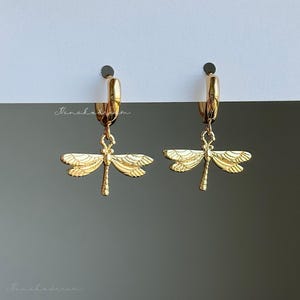 May include: A pair of gold-toned hoop earrings featuring dangling dragonfly charms. Each dragonfly pendant displays detailed wing textures and a slender body. The earrings are suspended from small, polished gold hoops, showcasing a metallic finish throughout the design.