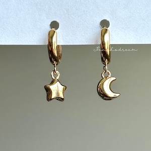 May include: A pair of gold-colored hoop earrings. One earring features a star charm, and the other has a crescent moon charm. The earrings are displayed against a neutral background, highlighting their celestial design.