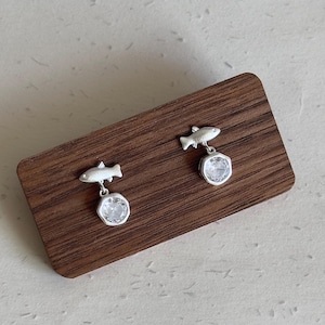 May include: A pair of silver earrings featuring a fish design above a clear, round gemstone. The earrings are displayed on a wooden surface, showcasing their delicate craftsmanship and aquatic theme.