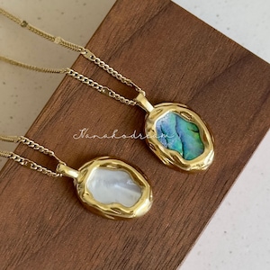 May include: Two gold-colored necklaces with oval pendants. One pendant features a white, pearlescent stone, and the other has a blue and green iridescent stone. The pendants have a textured, irregular gold border. The necklaces rest on a wooden surface.