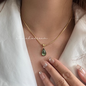 May include: A gold-colored necklace with a teardrop-shaped green pendant. The necklace is worn against a white shirt. The pendant is a light green color. The necklace is delicate and elegant.