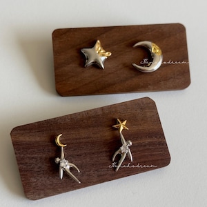 May include: Silver and gold-toned celestial earrings. One pair features a star and crescent moon, the other has a person reaching for a star. The earrings are displayed on small, rectangular wooden blocks.