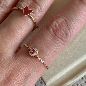 May include: Two gold rings on a hand. One ring has a red heart-shaped stone. The other ring has a square red stone surrounded by smaller clear stones.