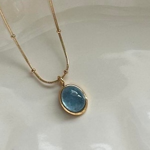 Peut inclure: Un collier dor&eacute; avec une cha&icirc;ne d&eacute;licate et un pendentif orn&eacute; d'une pierre pr&eacute;cieuse ovale bleu clair. Le pendentif est encadr&eacute; d'une lunette dor&eacute;e. Le collier est pr&eacute;sent&eacute; sur une surface blanche.