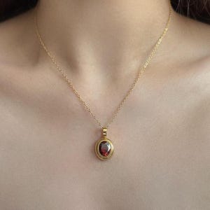 May include: A gold necklace with a pendant. The pendant has a red oval gemstone set in a gold frame, suspended from a fine gold chain. The necklace is shown against a neutral backdrop.