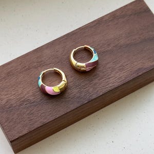 May include: A pair of gold-tone hoop earrings with colourful enamel detailing. Each earring features a segmented design with pink, yellow, teal, and purple enamel sections. They are presented on a dark brown wooden surface.