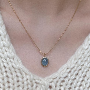 Peut inclure: Un collier dor&eacute; avec un petit pendentif ovale. Le pendentif contient une pierre bleu clair dans un cadre dor&eacute;. Le collier est port&eacute; sur un pull en tricot de couleur cr&egrave;me.