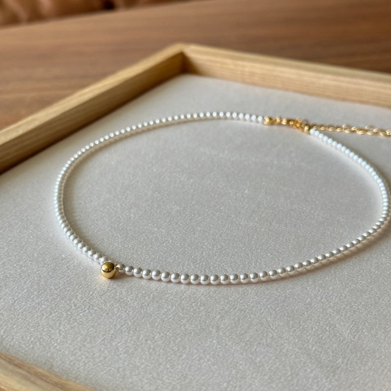 Peut inclure: Un collier d&eacute;licat de perles avec un petit accent de perle dor&eacute;e. Le collier est blanc et la perle est de couleur or brillant.