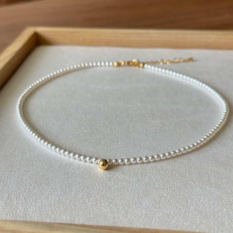 Peut inclure: Un collier d&eacute;licat de perles blanches avec un seul accent de perle dor&eacute;e. Le collier est pr&eacute;sent&eacute; dans une bo&icirc;te en bois.