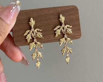 May include: A pair of gold-colored leaf-shaped earrings. Each earring features three cascading leaf clusters with small, clear gemstones. The earrings are displayed on a wooden surface against a neutral background.