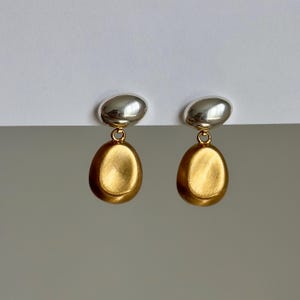 May include: A pair of silver and gold earrings. The earrings are shaped like a teardrop with a smooth, brushed finish. The top of the earring is a silver oval.