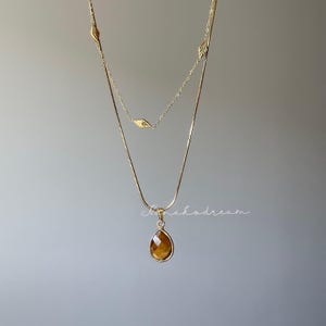 May include: A layered gold-toned necklace with a teardrop-shaped pendant. The pendant features a brown gemstone. The necklace has a delicate chain design with leaf-shaped accents. The background is a solid gray.