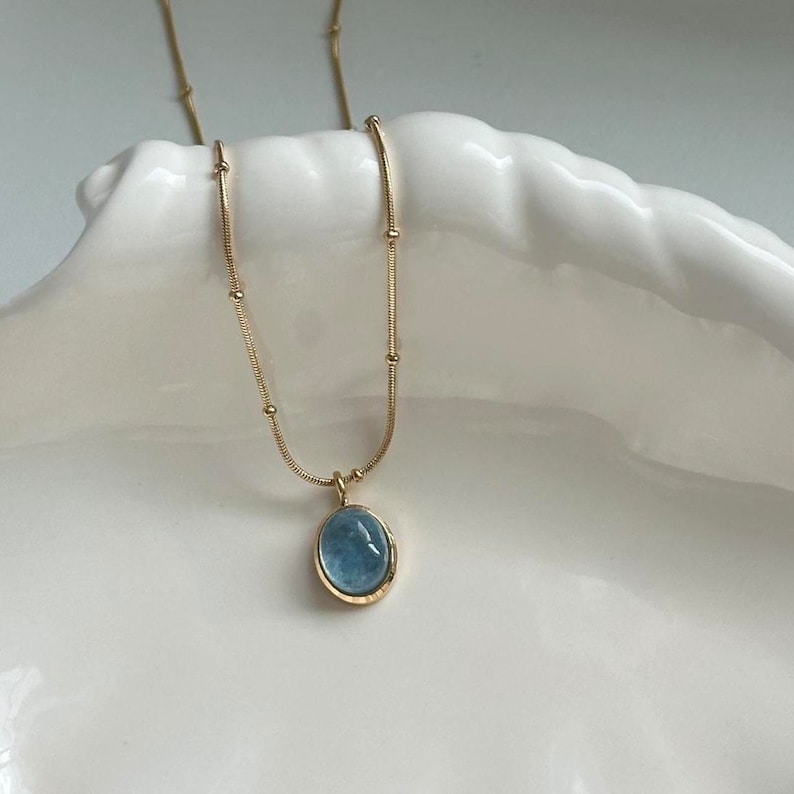 Peut inclure: Un collier en or d&eacute;licat avec un pendentif ovale bleu clair. Le pendentif est serti dans un cadre dor&eacute; et est suspendu &agrave; une fine cha&icirc;ne dor&eacute;e perl&eacute;e. Le collier est pr&eacute;sent&eacute; sur une surface blanche en forme de coquillage.