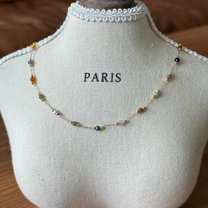 May include: A gold chain necklace with small, colourful beads. The beads are various shades of orange, red, green, white, and black. The necklace is displayed on a white mannequin bust with the word "PARIS" printed on the front.