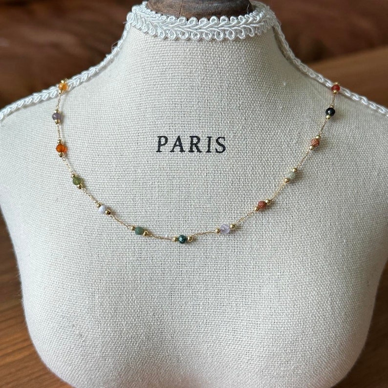 May include: A gold chain necklace with small, colourful beads. The beads are various shades of orange, red, green, white, and black. The necklace is displayed on a white mannequin bust with the word "PARIS" printed on the front.
