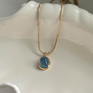 Peut inclure: Un collier dor&eacute; avec un pendentif ovale bleu clair. Le pendentif est serti dans un cadre dor&eacute; et est suspendu &agrave; une cha&icirc;ne d&eacute;licate. La cha&icirc;ne comporte de petites perles dor&eacute;es espac&eacute;es r&eacute;guli&egrave;rement. Le collier est pr&eacute;sent&eacute; sur une surface blanche.