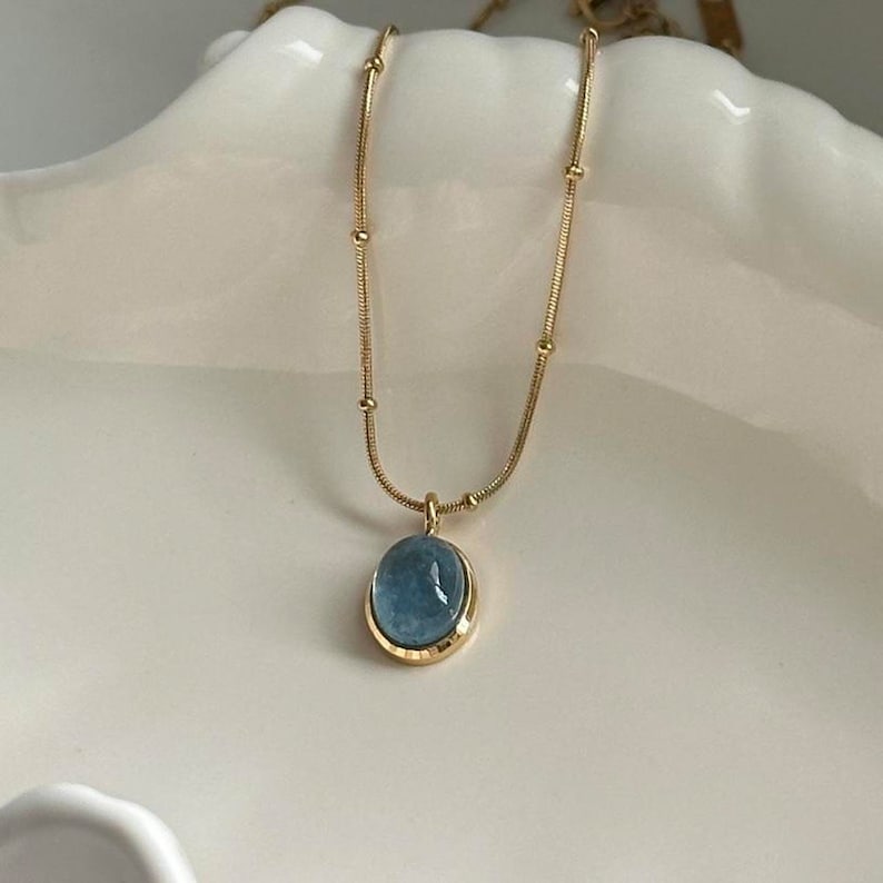 Peut inclure: Un collier dor&eacute; avec un pendentif ovale bleu clair. Le pendentif est serti dans un cadre dor&eacute; et est suspendu &agrave; une cha&icirc;ne d&eacute;licate. La cha&icirc;ne comporte de petites perles dor&eacute;es espac&eacute;es r&eacute;guli&egrave;rement. Le collier est pr&eacute;sent&eacute; sur une surface blanche.