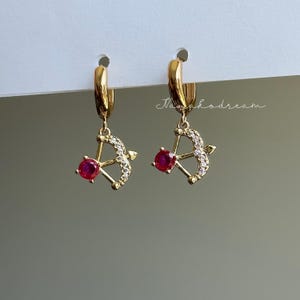 May include: A pair of gold-colored hoop earrings. Each earring features a small bow and arrow charm with a red gemstone at the center of the bow. The bow and arrow are encrusted with small, clear gemstones. The earrings are suspended from gold hoops.