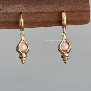 Peut inclure: Une paire de boucles d'oreilles créoles dorées avec une breloque en forme de coquillage en spirale. Chaque breloque est fabriquée en métal doré poli et contient une petite perle blanche ronde sertie au centre de la cavité du coquillage.