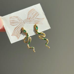 May include: Gold-colored snake earrings with green enamel detailing. Each earring features a serpentine design with a green gemstone accent at the head. The earrings are displayed on a white card with a bow illustration.