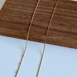 Peut inclure: Gros plan sur deux d&eacute;licats colliers dor&eacute;s. L'un des colliers pr&eacute;sente de petites perles espac&eacute;es r&eacute;guli&egrave;rement, tandis que l'autre est une cha&icirc;ne simple et fine. Les colliers sont pos&eacute;s sur une surface en bois et un fond blanc. Le texte "Nanahodream" est visible.