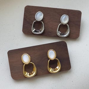 May include: Two pairs of earrings. One pair is silver-toned with a white oval stone and a teardrop-shaped design. The other pair is gold-toned with a white oval stone and a teardrop-shaped design. The earrings are displayed on wooden rectangles.