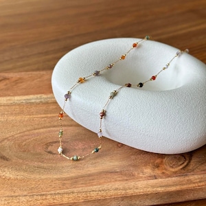 May include: A gold chain necklace with small, colourful gemstone beads. The necklace is displayed on a white, oval-shaped surface.