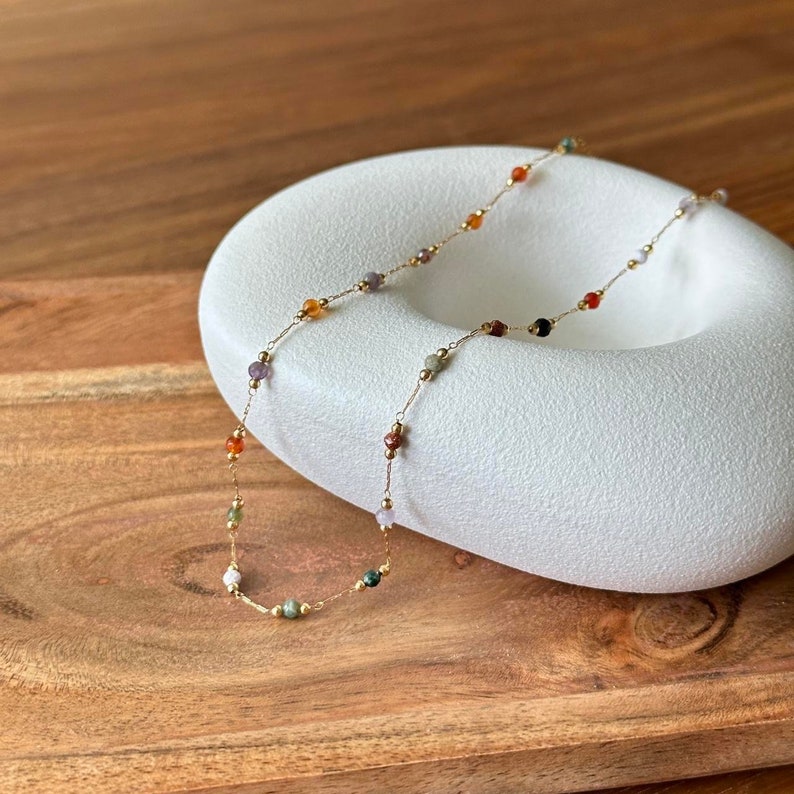 May include: A gold chain necklace with small, colourful gemstone beads. The necklace is displayed on a white, oval-shaped surface.
