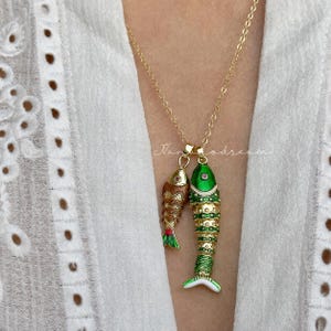 May include: A gold-tone chain necklace featuring two fish-shaped pendants. One pendant is smaller with brown and gold textured scales, while the larger pendant is green and gold with detailed scale patterns. Both fish have small red accents.