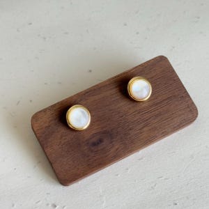 May include: Two gold-colored stud earrings with a white, pearlescent center are displayed on a dark brown wooden surface. The earrings have a simple, round design, and the gold setting frames the white center.